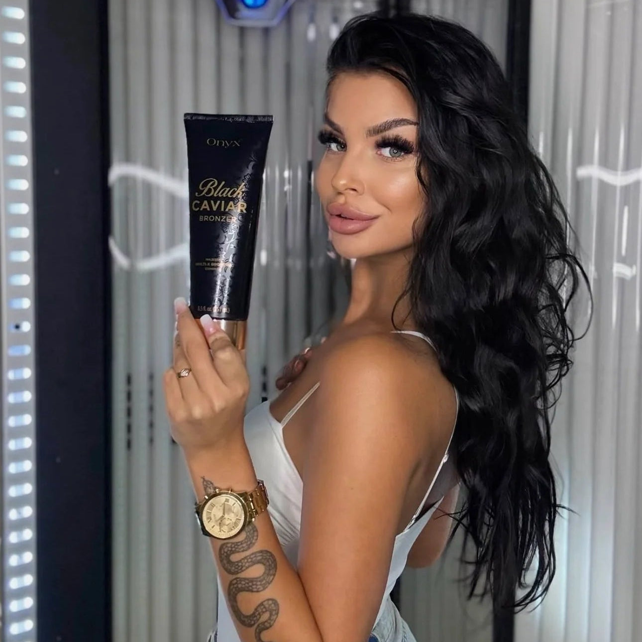 Model holding an Onyx Black Caviar Bronzer tube beside a sunbed in a tanning salon.