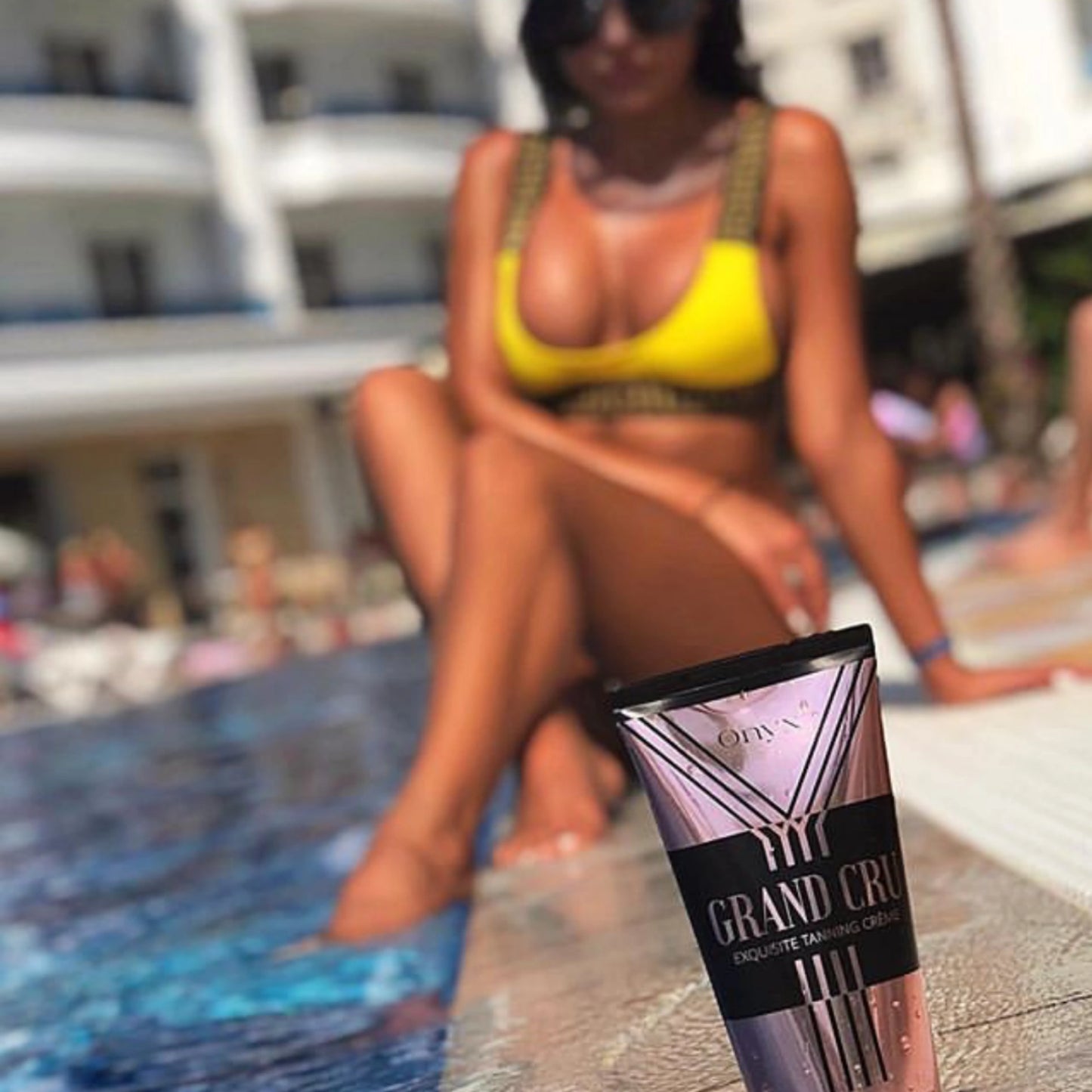 Onyx Grand Cru Tanning Accelerator placed poolside with a woman in the background, enjoying the sun in a yellow bikini.
