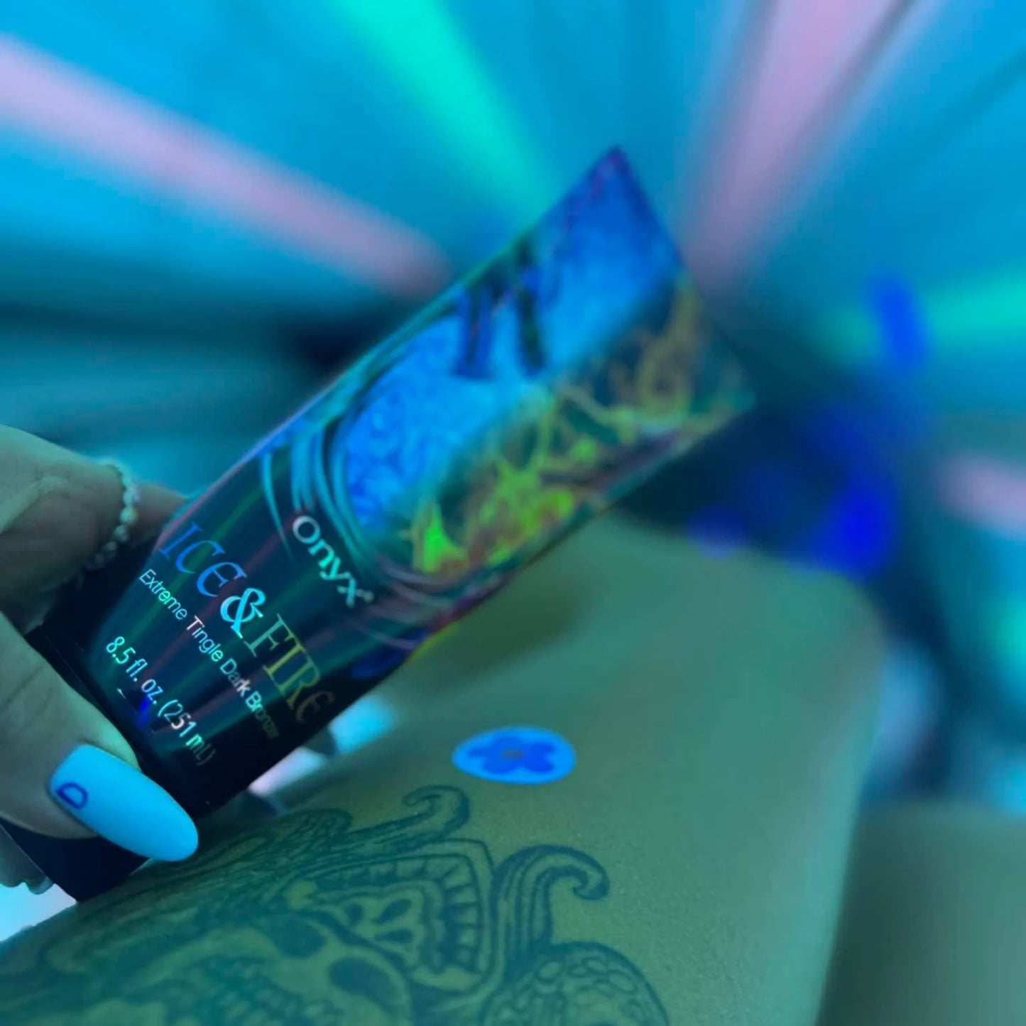 Hand holding Onyx Ice & Fire tanning lotion during blue-lit sunbed session, tattoo visible on the leg.
