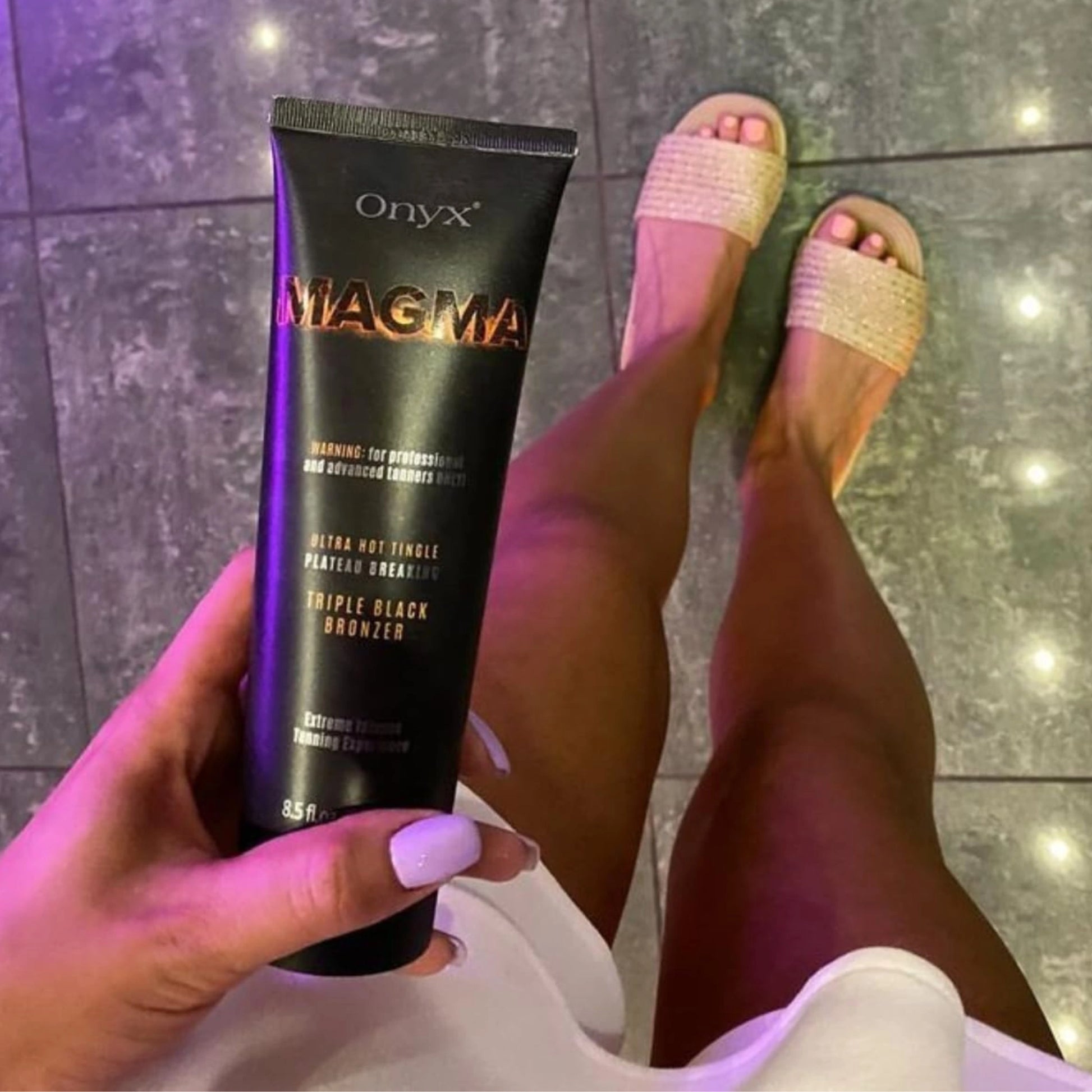 Woman holding Onyx Magma tingle tanning cream while showing her bronzed legs in white outfit and beige sandals on glossy floor