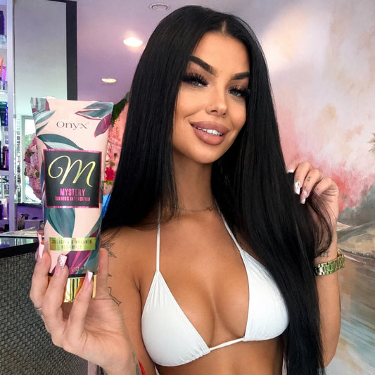Model holding an Onyx Mystery Tanning Intensifier cream indoors while wearing a white bikini.
