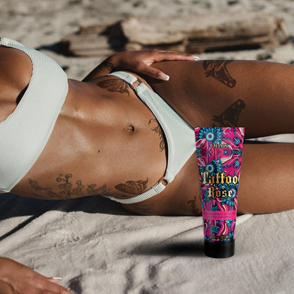 Woman with tattoos on stomach and legs lying on sandy beach wearing white bikini with Onyx Tattoo Rose tanning cream placed beside her