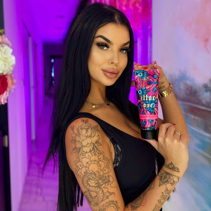 Woman with long black hair and dragon tattoo on her arm wearing black top and holding Onyx Tattoo Rose tanning cream in a purple-lit hallway