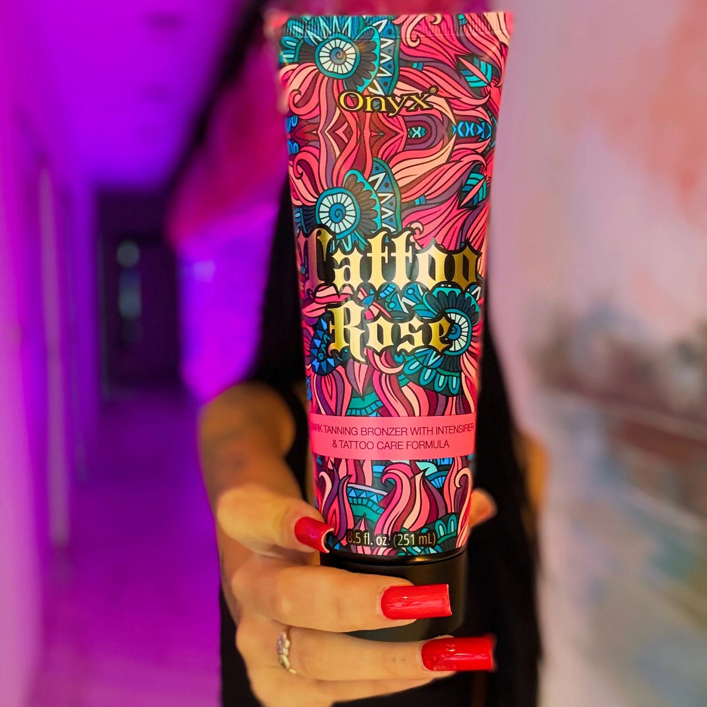 Woman with long red nails holding Onyx Tattoo Rose tanning cream toward camera in pink-lit hallway
