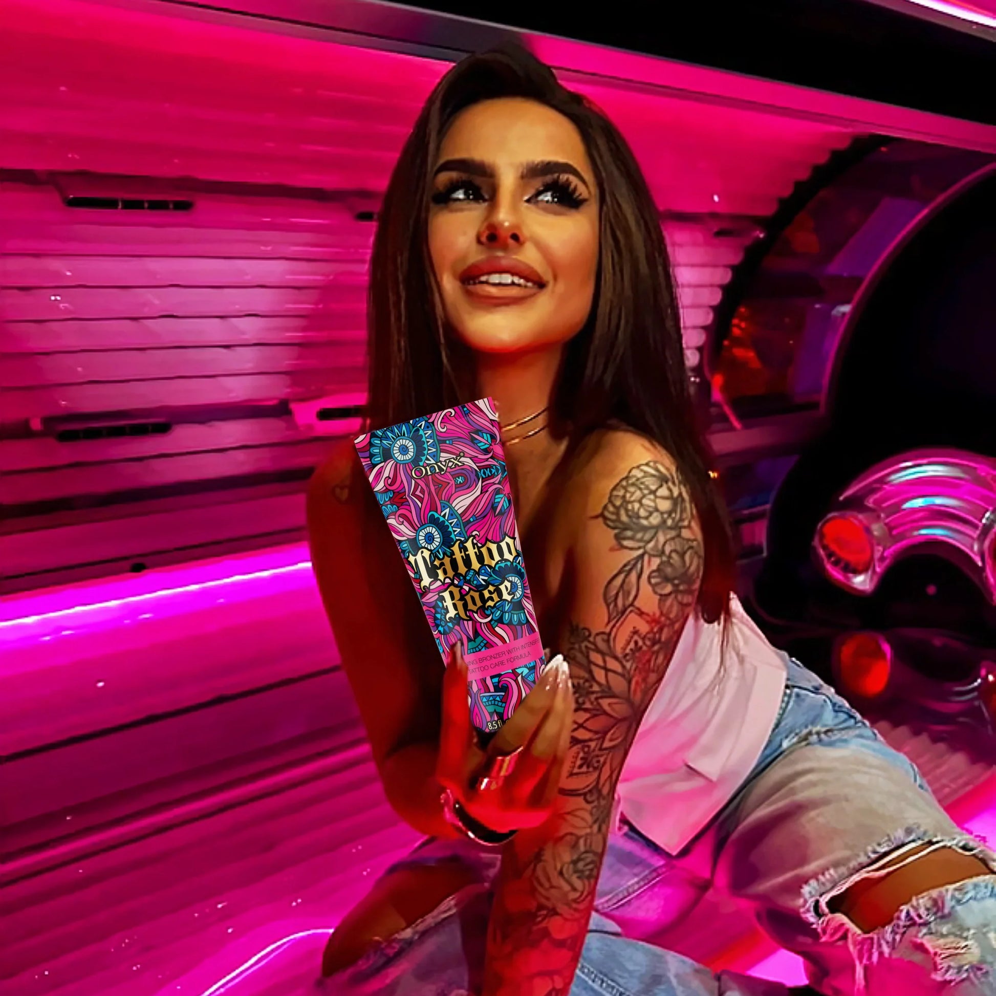 Smiling woman with long brown hair and floral tattoos posing near a tanning bed under bright pink light holding Onyx Tattoo Rose tanning cream