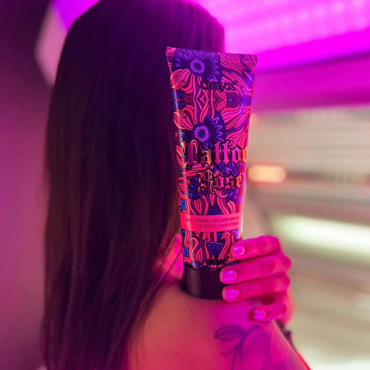 Woman showing shoulder tattoo holding Onyx Tattoo Rose tanning cream under purple light inside tanning room