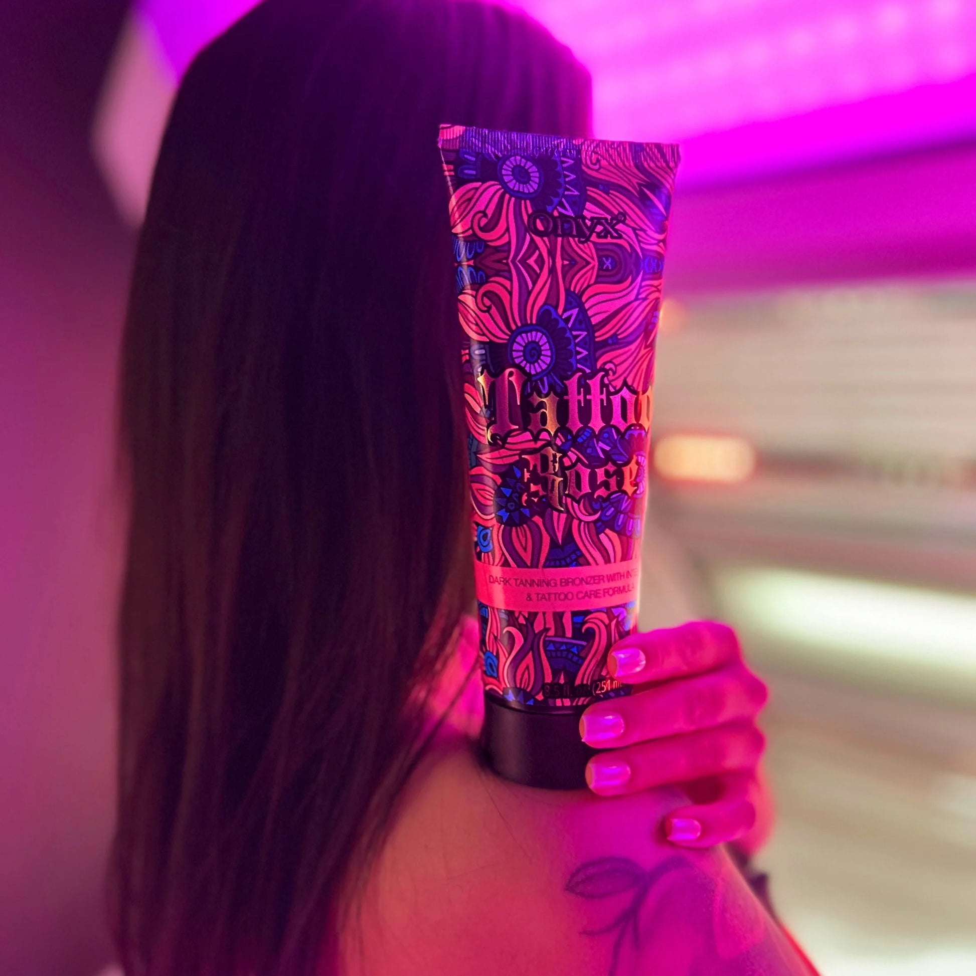 Woman showing shoulder tattoo holding Onyx Tattoo Rose tanning cream under purple light inside tanning room